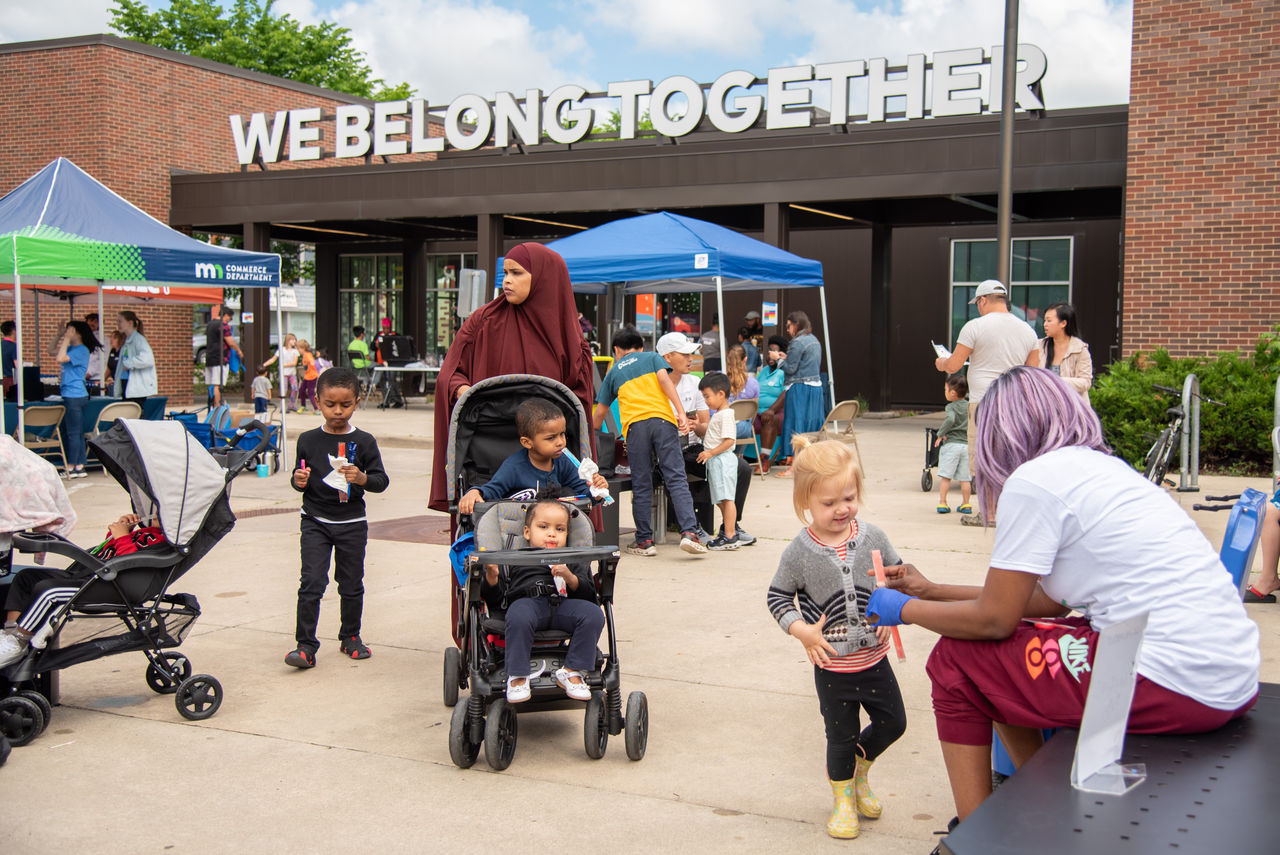 A woman in a hijab pushes a stroller in a crowd of people outside a community center.